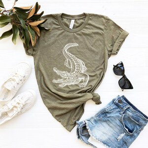 Puede incluir: Una camiseta verde con un gr&aacute;fico de un cocodrilo blanco. La camiseta est&aacute; colocada sobre una superficie blanca con un par de zapatillas blancas, unos pantalones cortos de mezclilla azul y unas gafas de sol negras.