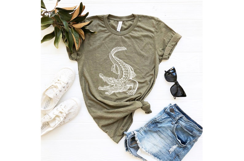Puede incluir: Una camiseta verde con un gr&aacute;fico de un cocodrilo blanco. La camiseta est&aacute; colocada sobre una superficie blanca con un par de zapatillas blancas, unos pantalones cortos de mezclilla azul y unas gafas de sol negras.