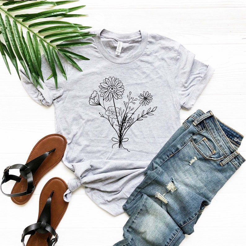 Pode incluir: Uma camiseta cinza com um desenho de linha preta de um buqu&ecirc; de flores. A camiseta est&aacute; colocada sobre uma superf&iacute;cie branca com um par de sand&aacute;lias marrons e cal&ccedil;as de ganga azuis.