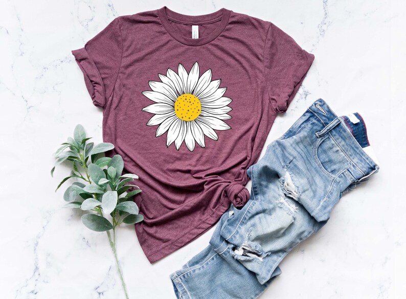 May include: A maroon t-shirt with a white daisy graphic. The daisy has a yellow center and is centered on the shirt. The shirt is laying on a white marble surface with a pair of blue jeans and a sprig of green foliage.