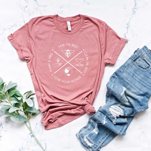 May include: A dusty rose t-shirt with a white graphic that reads "Save the Bees, Plant a Tree, Clean the Seas, Love Your Mother." The shirt is paired with light blue denim jeans with distressed details. A sprig of green leaves is in the corner.