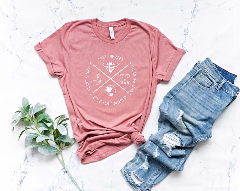 May include: A dusty rose t-shirt with a white graphic that reads "Save the Bees, Plant a Tree, Clean the Seas, Love Your Mother." The shirt is paired with light blue denim jeans with distressed details. A sprig of green leaves is in the corner.