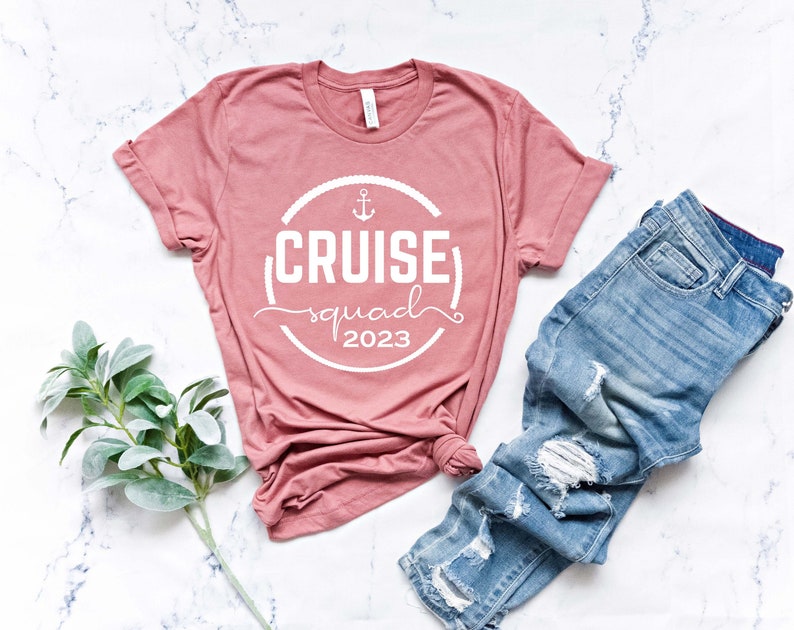 May include: A pink t-shirt with a white circle design that says "Cruise Squad 2023" with an anchor symbol. The shirt is laying on a white marble surface with a pair of blue jeans.