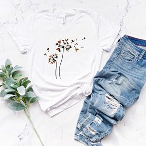 May include: White t-shirt with a graphic of three dandelion flowers with seeds blowing in the wind. The dandelion flowers are brown, orange, and green. The t-shirt is laying on a white marble surface with a pair of blue jeans and a sprig of green foliage.