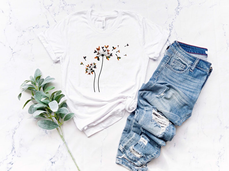 May include: White t-shirt with a graphic of three dandelion flowers with seeds blowing in the wind. The dandelion flowers are brown, orange, and green. The t-shirt is laying on a white marble surface with a pair of blue jeans and a sprig of green foliage.