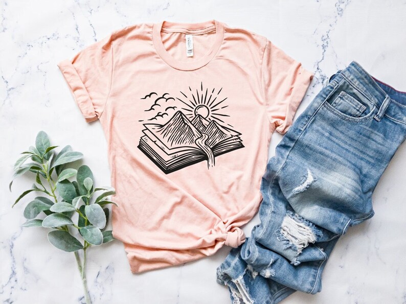 May include: A peach-colored t-shirt with a black and white graphic of a book with mountains and a sun rising above them.