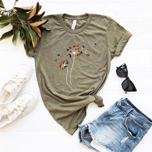 May include: A green t-shirt with a white dandelion graphic featuring colorful leaves. The dandelion is blowing seeds in the wind. The t-shirt is laid out on a white surface with a pair of white shoes, a pair of sunglasses, and a pair of blue denim shorts.