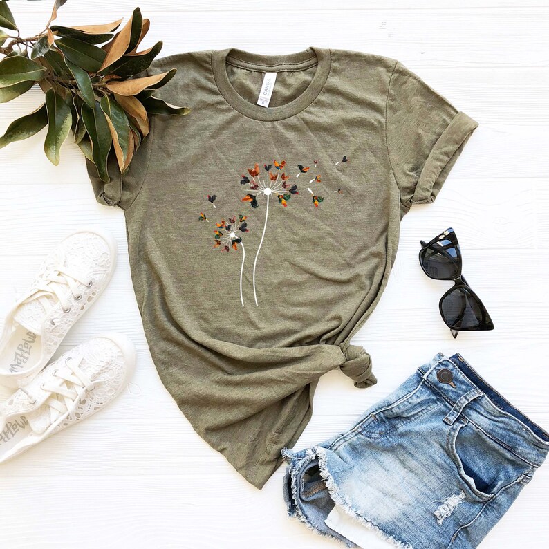 May include: A green t-shirt with a white dandelion graphic featuring colorful leaves. The dandelion is blowing seeds in the wind. The t-shirt is laid out on a white surface with a pair of white shoes, a pair of sunglasses, and a pair of blue denim shorts.