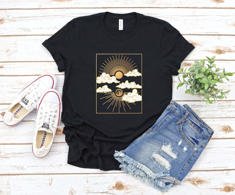May include: A black t-shirt with a gold graphic of a sun, moon, and clouds. The graphic is in a rectangular frame.