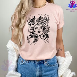 Pode incluir: Camiseta rosa claro com um desenho gr&aacute;fico em preto e branco do rosto da Medusa. O design mostra Medusa com cobras no cabelo, com tra&ccedil;os faciais detalhados. A camiseta tem mangas curtas e gola redonda.
