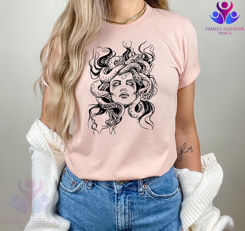 Pode incluir: Camiseta rosa claro com um desenho gr&aacute;fico em preto e branco do rosto da Medusa. O design mostra Medusa com cobras no cabelo, com tra&ccedil;os faciais detalhados. A camiseta tem mangas curtas e gola redonda.