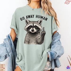 May include: A light green t-shirt with the text "GO AWAY HUMAN" above a detailed illustration of a raccoon giving the middle finger. The raccoon is in black and white, and the shirt is 100% cotton.
