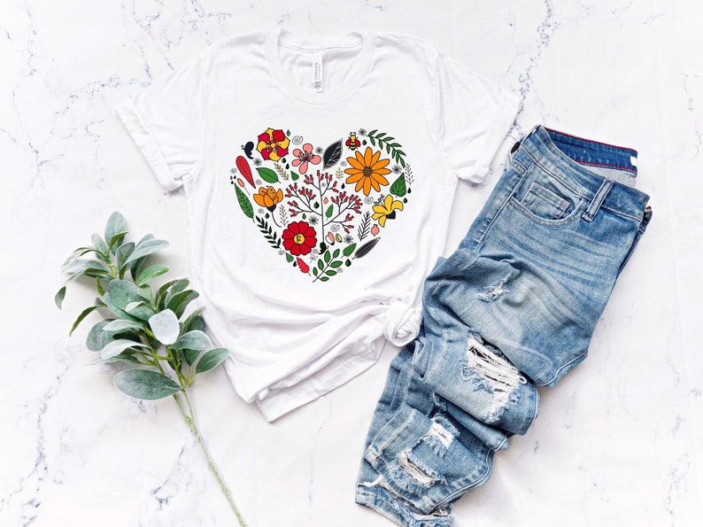 May include: White t-shirt with a floral heart design. The heart is made up of various flowers, leaves, and other botanical elements in shades of red, orange, yellow, green, and brown.