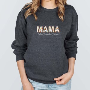 May include: A dark gray sweatshirt with the word "MAMA" printed in colorful letters. The letters are decorated with a floral pattern. Below the word "MAMA" is the text "Robin Jennifer Emma" with a heart symbol between each name.