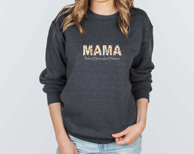 May include: A dark gray sweatshirt with the word "MAMA" printed in colorful letters. The letters are decorated with a floral pattern. Below the word "MAMA" is the text "Robin Jennifer Emma" with a heart symbol between each name.