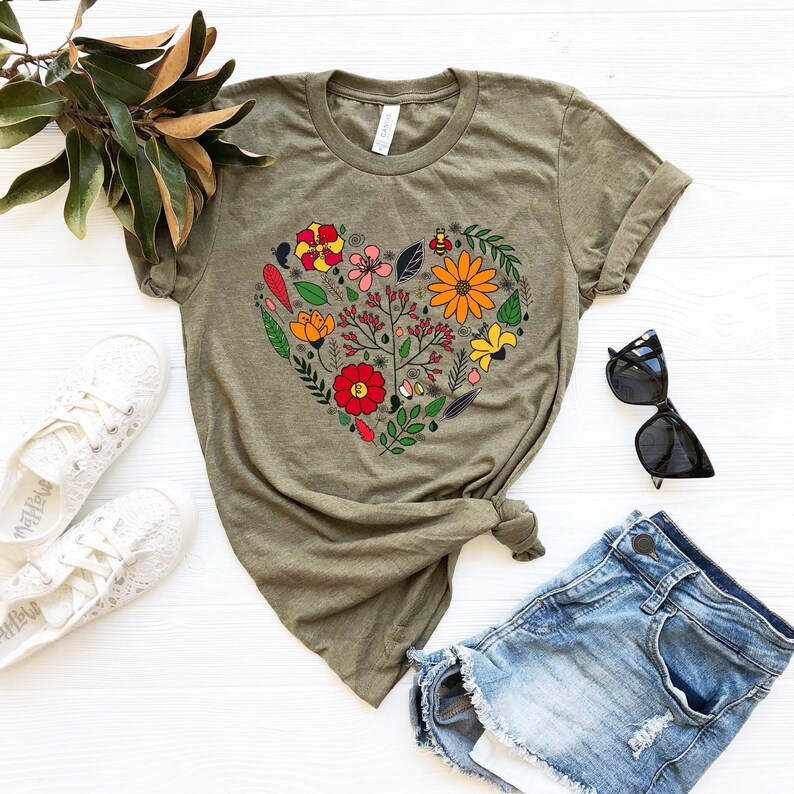 May include: A green t-shirt with a floral heart design. The heart is made up of various flowers, leaves, and a bee. The shirt is laid out on a white surface with a pair of white sneakers, a pair of sunglasses, and a pair of denim shorts.
