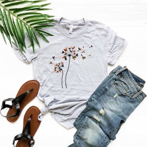 May include: A heather grey t-shirt with a graphic of a dandelion with colorful leaves blowing in the wind. The shirt is paired with blue jeans and brown sandals.