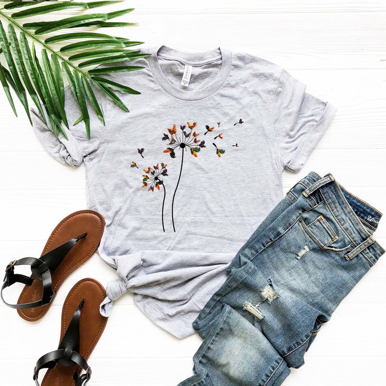 May include: A heather grey t-shirt with a graphic of a dandelion with colorful leaves blowing in the wind. The shirt is paired with blue jeans and brown sandals.