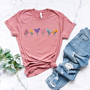 May include: A dusty rose t-shirt with a row of colourful flowers printed on the front. The shirt is made of a soft, comfortable material and has a relaxed fit.