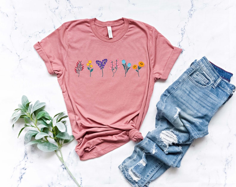 May include: A dusty rose t-shirt with a row of colourful flowers printed on the front. The shirt is made of a soft, comfortable material and has a relaxed fit.