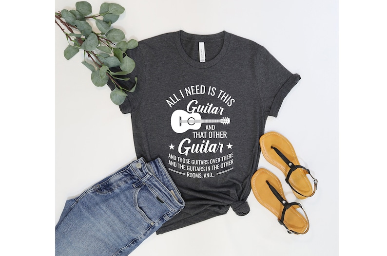 Puede incluir: Una camiseta gris oscuro con un gr&aacute;fico blanco que dice "Todo lo que necesito es esta guitarra y esa otra guitarra" con un dibujo de una guitarra. El texto contin&uacute;a con "y esas guitarras de all&aacute; y las guitarras en las otras habitaciones, y..."