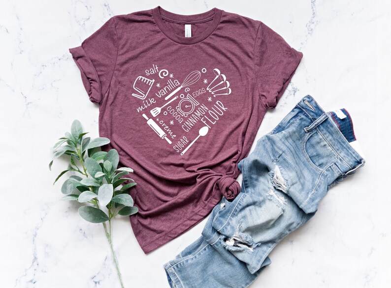 Może przedstawiać: Bordowy t-shirt z białym nadrukiem przedstawiającym składniki i narzędzia do pieczenia. Nadruk zawiera słowa "s&oacute;l", "wanilia", "mleko", "jajka", "kakao", "śmietanka", "cukier", "cynamon" i "mąka".