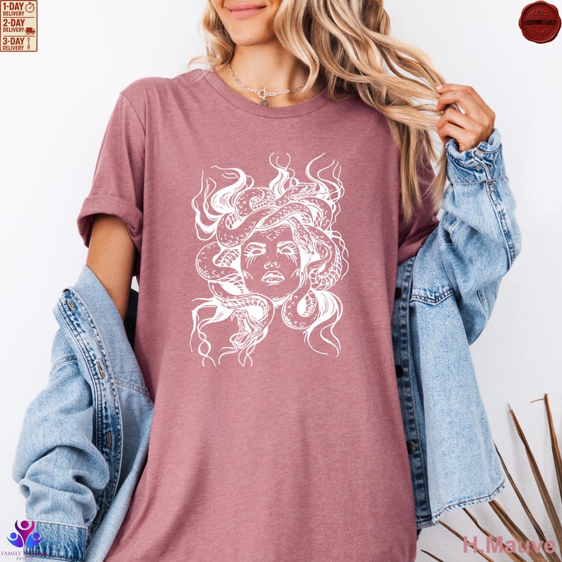 Pode incluir: Camiseta cor de malva com um gr&aacute;fico branco do rosto de Medusa e cobras. O design &eacute; um desenho de linha detalhado. A camisa tem mangas curtas e um ajuste folgado. A modelo est&aacute; usando uma jaqueta jeans.