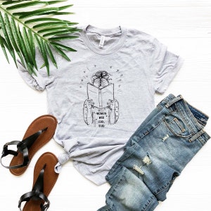 May include: A light gray t-shirt with a black graphic of a person reading a book, with the text "WOMEN WHO LEAD, READ." The shirt is paired with blue jeans and brown sandals. A green leaf is in the background.