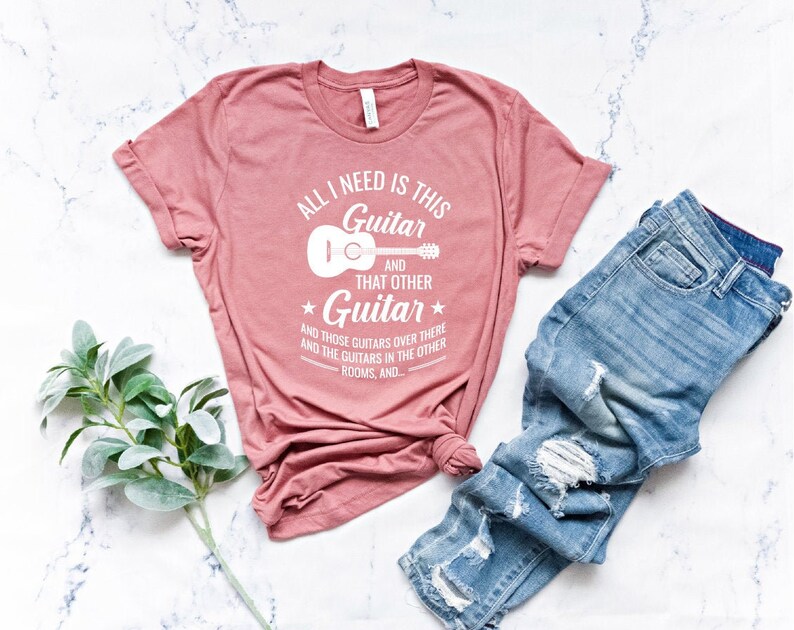 Puede incluir: Una camiseta rosa con el texto "All I need is this Guitar and that other Guitar" y un peque&ntilde;o gr&aacute;fico de guitarra. El texto contin&uacute;a con "and those guitars over there and the guitars in the other rooms, and..."