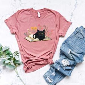 Puede incluir: Una camiseta rosa con un gato negro sentado en un libro abierto. El libro est&aacute; rodeado de peque&ntilde;as flores y hojas.