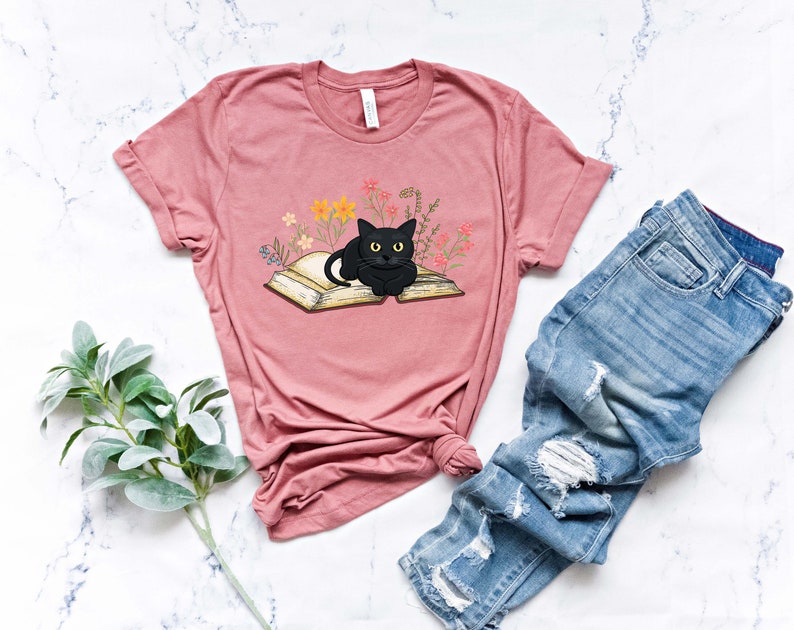 Puede incluir: Una camiseta rosa con un gato negro sentado en un libro abierto. El libro est&aacute; rodeado de peque&ntilde;as flores y hojas.