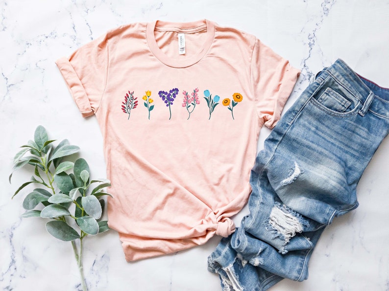 May include: A light pink t-shirt with a row of colourful flowers printed on the front. The flowers are in a variety of colours, including red, yellow, blue, purple, and green.