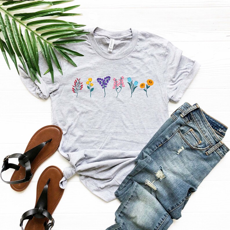 May include: A heather grey t-shirt with a row of colourful flowers printed on the front. The shirt is laid out on a white surface with a pair of brown sandals and blue jeans.