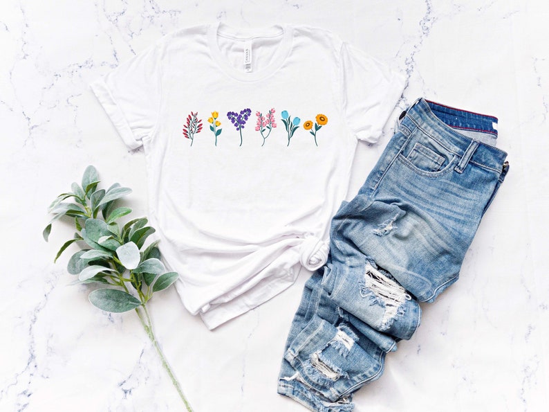 May include: White t-shirt with a row of colourful flowers printed on the front. The shirt is lying flat on a white marble surface with a pair of blue jeans and a sprig of greenery.