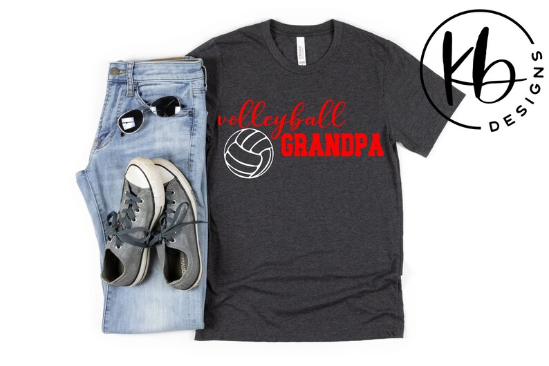 K&ouml;nnte beinhalten: Ein dunkelgraues T-Shirt mit dem Text "Volleyball Grandpa" in roten Buchstaben und einer wei&szlig;en Volleyballgrafik.