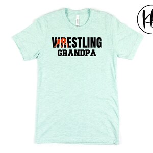 Puede incluir: Una camiseta azul claro con el texto "Wrestling Grandpa" en letras negras. Una silueta roja de un luchador est&aacute; incorporada en la palabra "Wrestling".