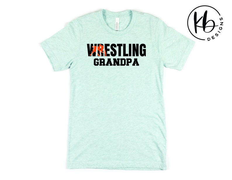 Puede incluir: Una camiseta azul claro con el texto "Wrestling Grandpa" en letras negras. Una silueta roja de un luchador est&aacute; incorporada en la palabra "Wrestling".