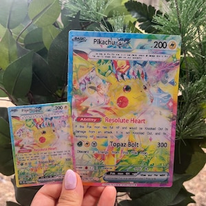 Puede incluir: Dos cartas coleccionables Pikachu EX. La carta más grande presenta un Pikachu amarillo con acentos coloridos y el texto "Resolute Heart" y "Topaz Bolt". La carta más pequeña refleja el diseño. Ambas cartas tienen una clasificación de 200 HP.