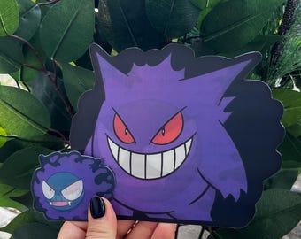 May include: Two purple and black Ghost Pokemon stickers. The larger sticker features a larger Ghost Pokemon with a wide, toothy grin. The smaller sticker features a smaller Ghost Pokemon with a smaller grin.
