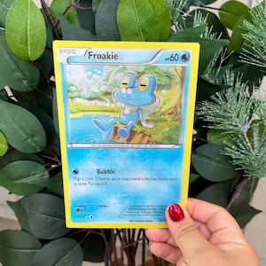 May include: A blue and white Pokemon trading card featuring Froakie, a blue frog Pokemon with a white belly and large, round eyes. The card has a yellow border and the text "Froakie" in black letters. The card also has a blue and white background with a picture of Froakie sitting on a log in a forest.