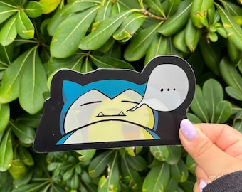 May include: A black sticker featuring a cartoon Snorlax character with closed eyes and a speech bubble. The Snorlax has blue, yellow, and black accents. The sticker is held in front of a background of green leaves.