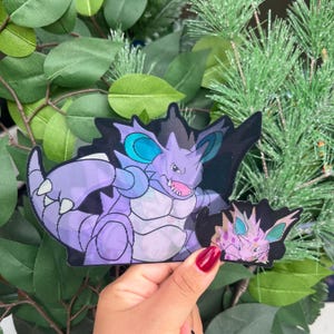 May include: A die-cut vinyl sticker featuring a large purple creature with teal ears, sharp teeth, and a spiked tail, accompanied by a smaller matching sticker of the same creature in a pink and purple color palette with teal accents.