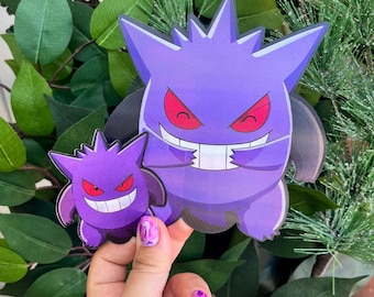 May include: Two purple Gengar character cutouts with red eyes and white teeth. The larger Gengar has arms crossed and a mischievous grin. The smaller Gengar has a similar design. The cutouts are set against a backdrop of green foliage.