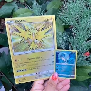 May include: A large yellow Zapdos trading card with 120 HP and a smaller blue Articuno trading card with 120 HP. The Zapdos card features electric yellow artwork, while the Articuno card displays blue icy imagery. Both cards show text.