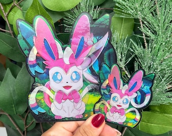 May include: Two holographic stickers featuring the character Sylveon, a white and pink creature with large blue eyes and ribbon-like feelers. The stickers have a glossy, reflective finish and display a vibrant, colorful forest background scene.