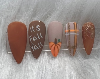 May include: A set of five fall-themed press-on nails. The nails feature various designs, including solid brown, the text "It's Fall Y'all", a pumpkin, a plaid pattern, and glitter. The nails are almond-shaped and are displayed on a clear plastic holder.