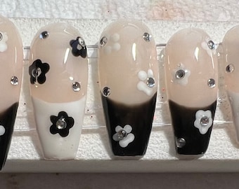 Könnte beinhalten: Set mit zehn langen, mandelförmigen falschen Nägeln mit einem Schwarz-Weiß-Ombre-Design. Jeder Nagel ist mit kleinen weißen und schwarzen Blumen und Strasssteinen verziert.