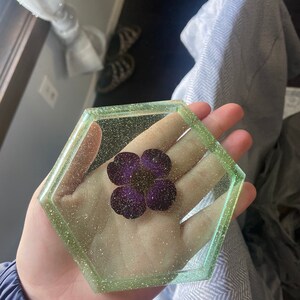 Puede incluir: Un posavasos hexagonal de resina con un borde verde brillante y una flor morada prensada en el interior.