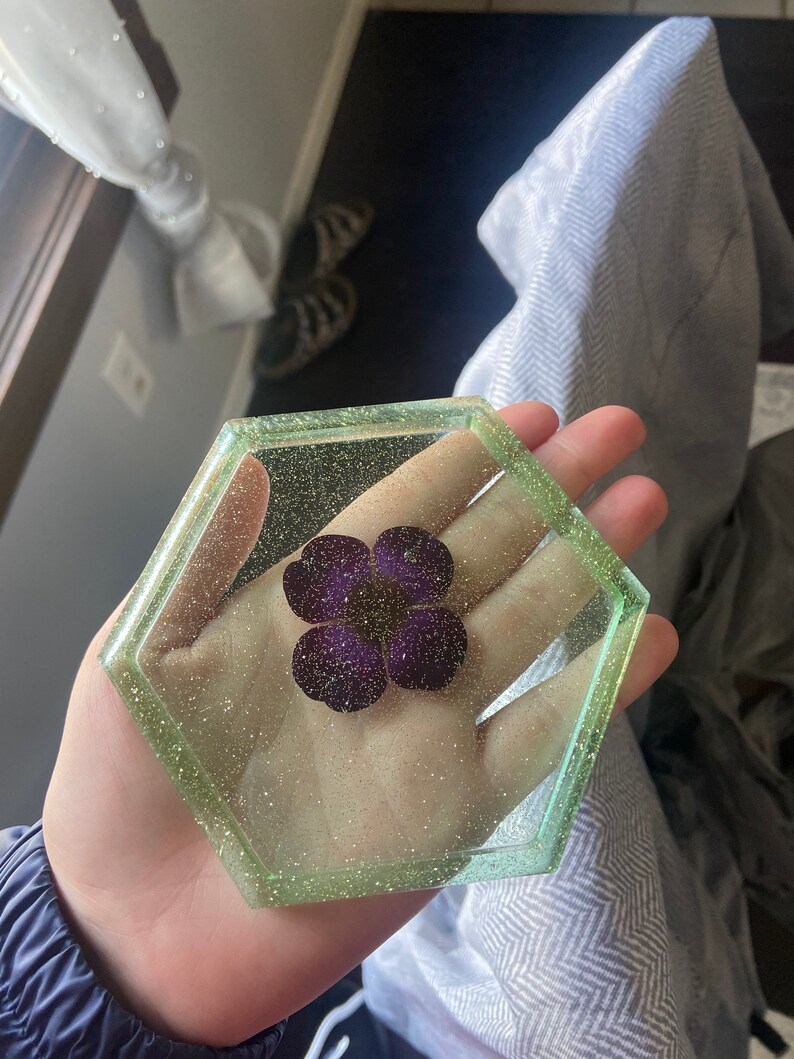 Puede incluir: Un posavasos hexagonal de resina con un borde verde brillante y una flor morada prensada en el interior.