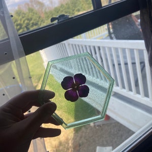 Puede incluir: Un posavasos hexagonal de resina verde y blanca con una flor morada prensada en el centro.